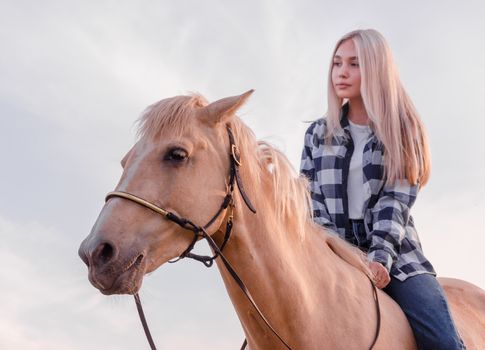 young blonde girl dressed in blue jeans and a plaid shirt sits on a horse at the ranch closeup