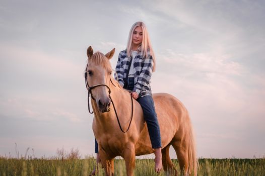 young blonde girl dressed in blue jeans and a plaid shirt sits on a horse at the ranch closeup