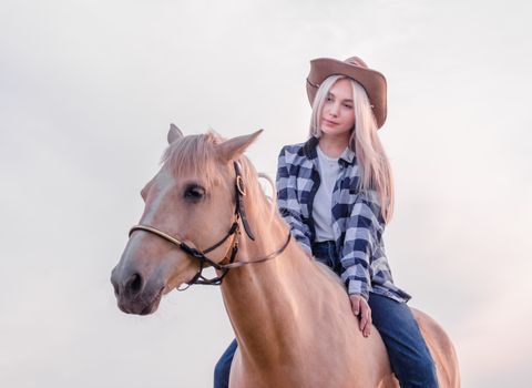 young girl dressed in a cowboy hat and blue jeans sits on a beige horse