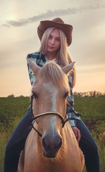 young girl dressed in a cowboy hat and blue jeans sits on a horse against the backdrop of the sunset