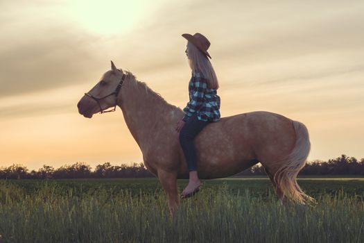 young girl dressed in a cowboy hat and blue jeans sits on a horse against the backdrop of the sunset