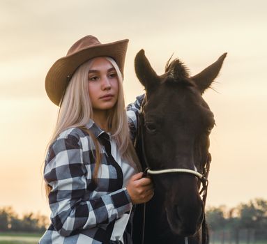 young girl dressed in a cowboy hat and a plaid shirt with a horse at sunset