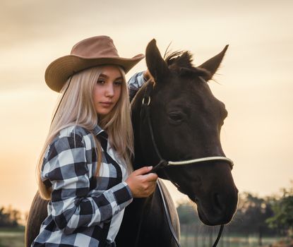 young girl dressed in a cowboy hat and a plaid shirt with a horse at sunset