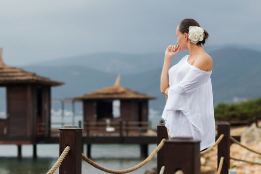 Lovely young female in stylish white blouse keeping eyes closed while standing on background of wonderful resort
