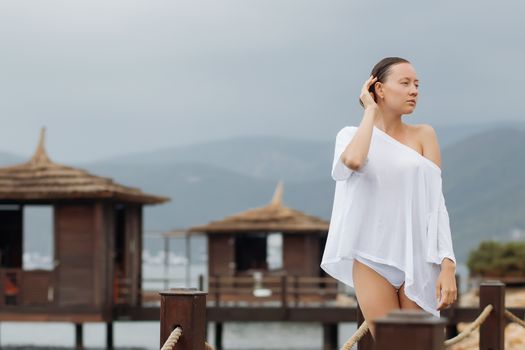 Lovely young female in stylish white blouse keeping eyes closed while standing on background of wonderful resort