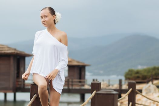 Lovely young female in stylish white blouse keeping eyes closed while standing on background of wonderful resort