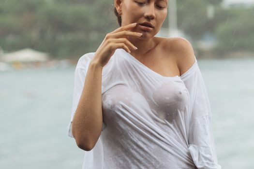 Attractive young female in while blouse and panties embracing herself and keeping eyes closed while standing near water on windy day