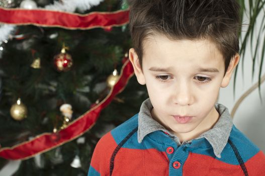 Unhappy little boy on christmass. Christmas tree in the background