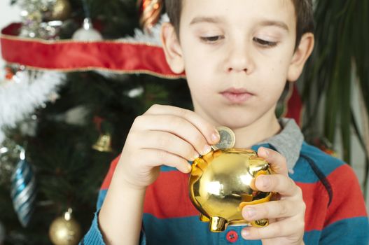 Little boy puts a coin in cash pig in front of the Christmass tree.