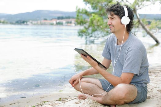 young man holding a tablet with headphones, at the beach