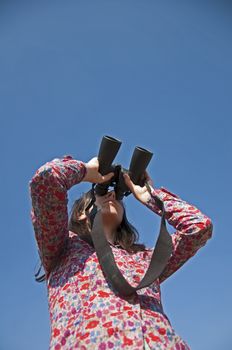 Young woman using binoculars. On blue sky background