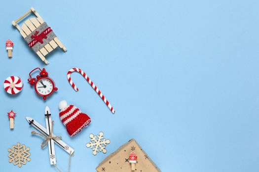 Corner creative Christmas composition. Candy cane, gift in craft paper, sled with deer, hat, alarm clock, ski, clothespins, wooden snowflakes on blue background, copy space. Minimal style. Top view.
