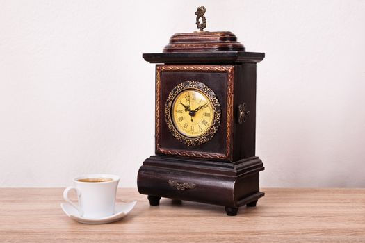 Time for coffee concept. Hot cup of coffee next to an old fashioned antique clock on a wooden table.