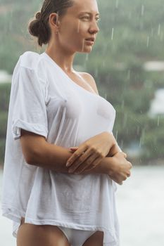 Attractive young female in while blouse and panties embracing herself and keeping eyes closed while standing near water on windy day