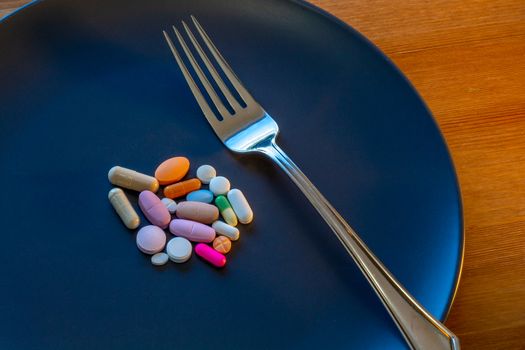 A dark gray plate has an assortment of pills at its center, including capsules and tablets from prescription medications, over-the-counter drugs, vitamins and other supplements. A fork is beside them.