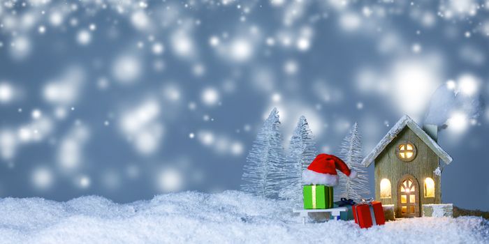 Christmas card with small glowing decorative house , gifts and Santa Claus cap on sleigh on snow over fairy tale winter fir forest background and falling snow . New Year design postcard . Copy space for text