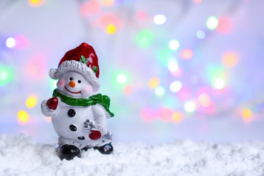 Smiling happy ceramic snowman in the snow, on a background of blurred christmas garland. Colorful festive bokeh effect. Space for the copy.