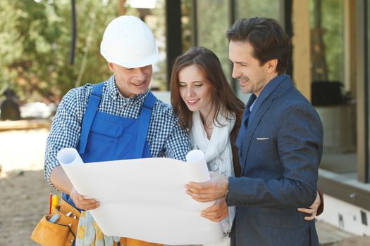 Foreman shows house design plans to a young couple at construction site outdoors