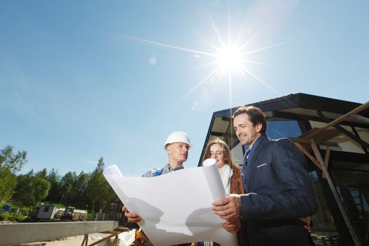 Foreman shows house design plans to a young couple at construction site outdoors at sunny day