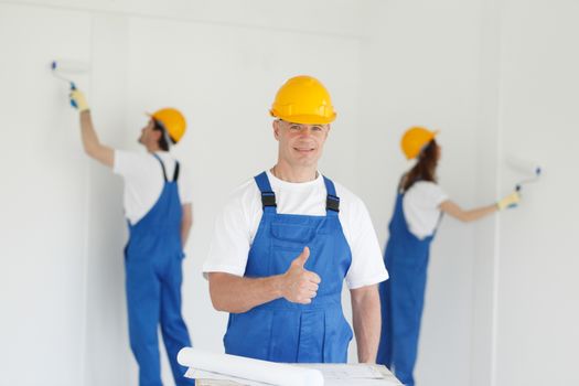 Manual worker gives thumbs up in front of two house painters painting walls