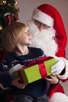 Portrait of smiling little boy sitting on santa claus knees and getting a gift