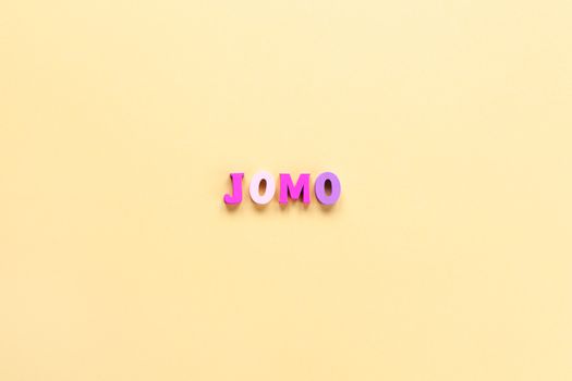 Abbreviation word JOMO in multicolored wooden letters on pastel yellow background. JOMO - Joy Of Missing Out. Opposition, choice, social problem, digital detox. Flat lay, copy space, minimalism style.