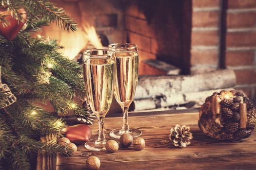 Christmas composition - Two glasses with champagne on a wooden table near a Christmas tree in a room with a burning fireplace.