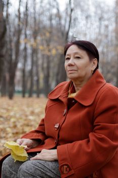 An elderly woman in a bright terracotta coat sits thoughtfully in an autumn Park