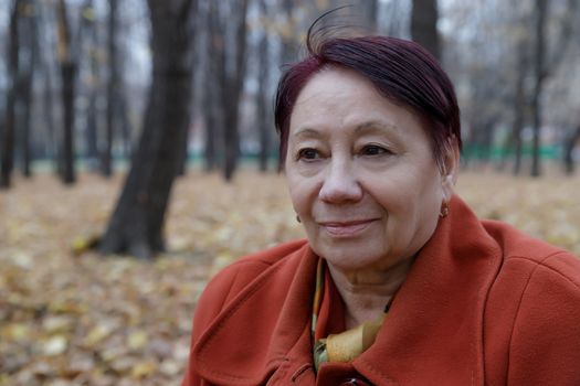 An elderly woman in a bright terracotta coat sits thoughtfully in an autumn Park