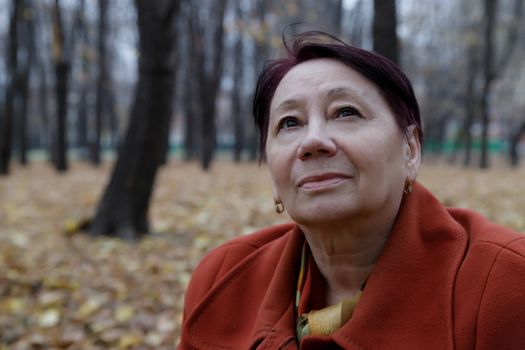 An elderly woman in a bright terracotta coat sits thoughtfully in an autumn Park