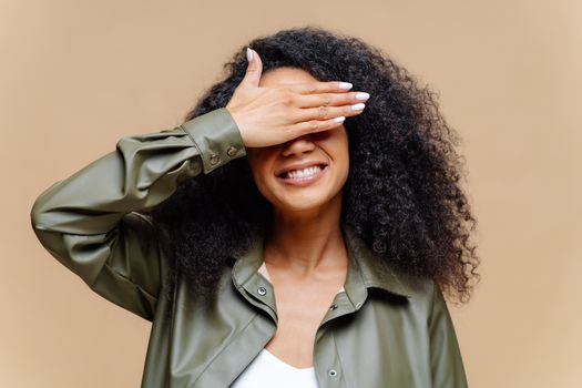 Shy cheerful young Afro woman covers eyes with palm, has toothy smile, hides face, has curly hairstyle, dressed in stylish clothes, isolated over brown background, waits for surprise or gift