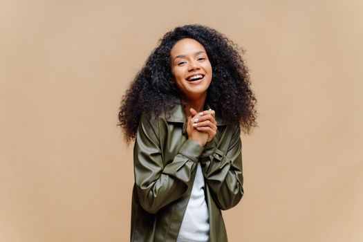 Positive pretty Afro American woman has curly hair, keeps hands together, happy to meet friend, dressed in fashionable clothes, smiles broadly, models against brown background. Wow, thats nice