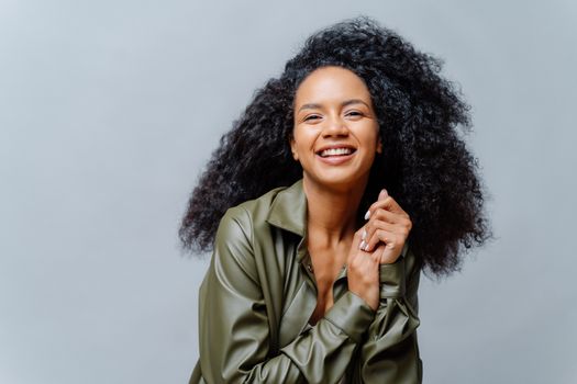 Joyful woman with dark skin, Afro hairstyle, keeps hands together, smiles sincerely at camera, shows white teeth, wears leather shirt, poses over grey background. People, emotions, ethnicity