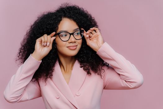 Headshot of attentive female boss keeps hands on frame of glasses, looks pensively away, wears rosy formal suit, has curly Afro hairstyle, poses against purple background. People, business, ethnicty