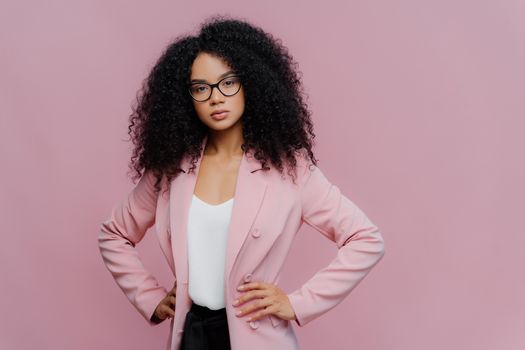 Photo of serious curly haired woman looks confidently at camera, wears formal neat clothes, keeps both hands on waist, wears optical glasses, poses over purple background. People, style concept