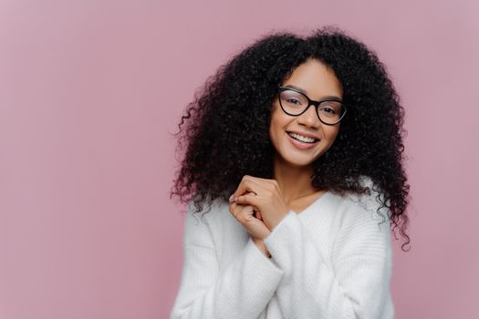 Portrait of happy positive African American woman with curly bushy hair, keeps hands together, has pleased facial expression, wears spectacles and white sweater, poses over purple background