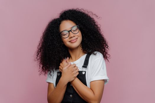 Portrait of satisfied woman with Afro hairstyle, keeps hands pressed on chest, tilts head, wears white casual t shirt and overalls, poses over purple background, recalls pleasant moment in life.