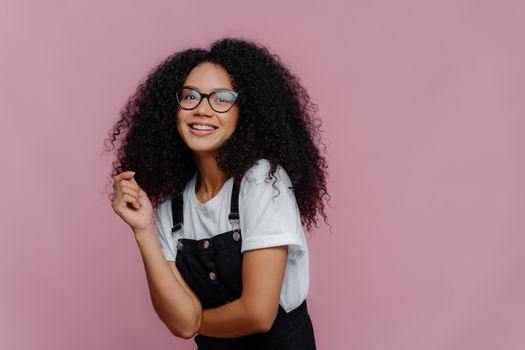 Studio shot of happy dark skinned lady with crisp hair, has dreamy expression, wears optical glasses, stylish clothes, imagines something pleasant, stands against bright violet background, free space