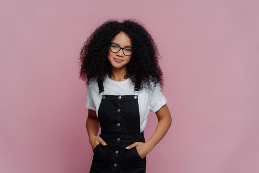Portrait of happy curly African American woman keeps hands in pockets of sarafan, wears transparent glasses, meets with friends during weekend, models over violet background, has delighted look
