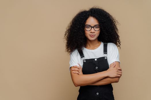 Photo of beautiful dark skinned teenage girl poses with arms crossed, wears spectacles, white t shirt and black sarafan, stands against brown background, copy space for your promotional content
