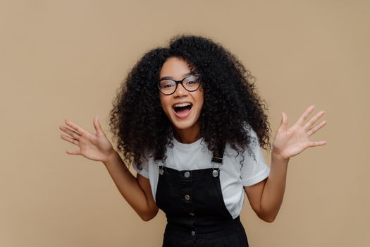 Overjoyed African American woman laughs out, raises palms, being in high spirit, wears transparent glasses, casual t shirt and overalls, poses against brown background, feels happiness and pleasure