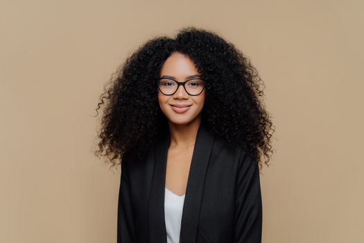 Portrait of beautiful Afro American woman with crisp hair, dressed in elegant black jacket, transparent glasses, looks directly at camera with gentle smile wears optical glasses isolated on brown wall