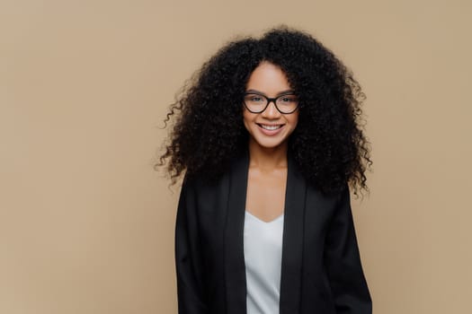 Portrait of glad curly haired young businesswoman with toothy smile, has delighted expression, wears spectacles and formal clothes, ready for meeting, poses over brown background. Lady in black