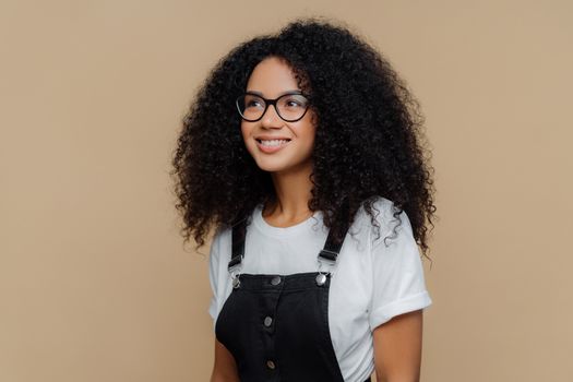 Pleased dreamy woman with curly hairstyle, looks through transparent glasses, dressed in casual white t shirt and black dungarees, poses against brown background, has good mood. People and emotions