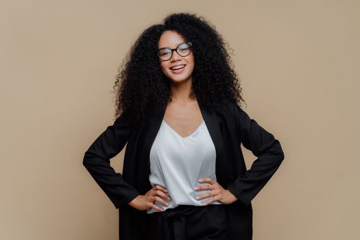 Half length shot of happy Afro woman dressed in elegant black clothes, keeps both hands on hips, wears optical glasses, smiles broadly, isolated over brown background. Female director in good mood