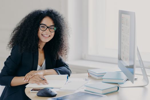 Glad curly office worker watches webinar on business topic, writes down notes with pen, smiles positively, wears spectacles and black clothes prepares report wears transparent glasses. Coworking space