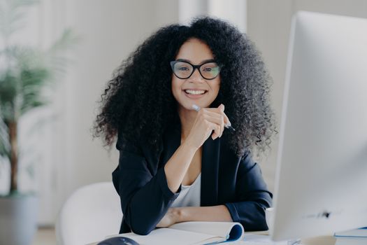 Positive international female student with crisp hair, wears transparent glasses, holds pen in hand, makes accountings, sits in front of big computer screen, dressed in black elegant outfit.