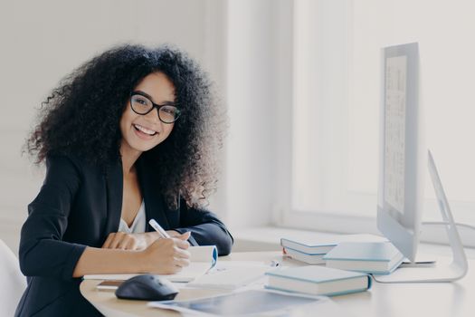 Cheerful female student writes down necessary information in notebook, makes notes, has glad expression, wears transparent glasses and black formal wear, sits at desktop with books and computer