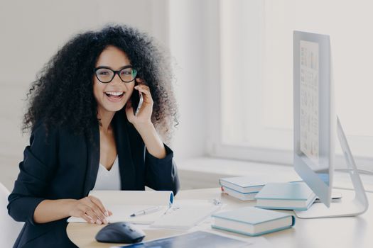 Optimistic female Afro American entrepreneur with curly hairstyle, holds mobile phone, enjoys telephone conversation with colleague, works on computer, poses at workplace with notepad and books