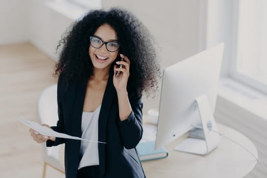 Cheerful female business worker calls partner to discuss details of statrup project, holds paper documents, wears optical glasses and elegant suit, gets necessary information, dressed elegantly